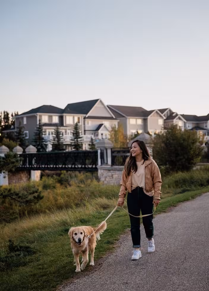 Woman walking a golden retriever on a Griesbach neighbourhood trail near a bridge, lakeside grasses, and nearby homes.