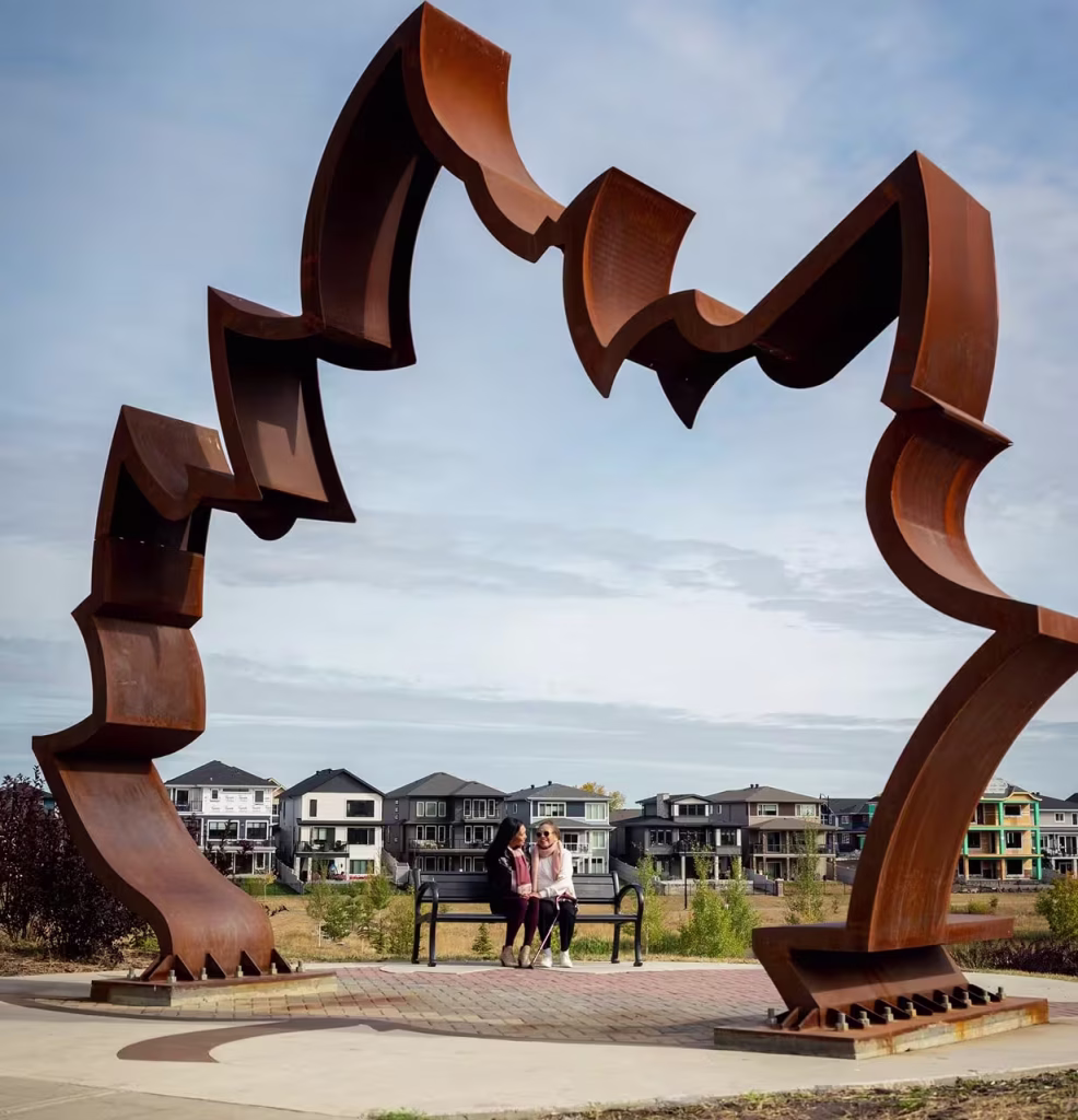 Une grande sculpture de Griesbach, de couleur rouille, encadre deux personnes assises sur un banc dans un parc résidentiel.